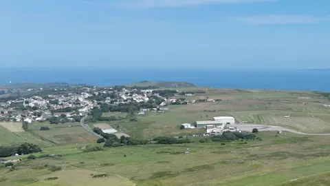 A picture taken from above looking down across Alderney. It is an island with a lot of grassland and houses in the distance. 