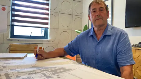 BBC Peter seated at a conference table in an office, holding a pen over a large architectural site plan spread across the tabletop. The room features a window with striped blinds, textured wall panels with circular patterns, framed artwork, and a wall-mounted television. A notebook and mug sit nearby, and the architectural drawings show a detailed layout of buildings, roads, and parking areas.