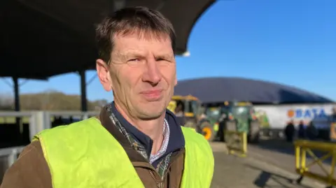 Stephen Ridsdale is wearing a hi vis jacket over the top of a brown waxed jacket. He has short brown hair and is standing in a yard containing agricultural vehicles.