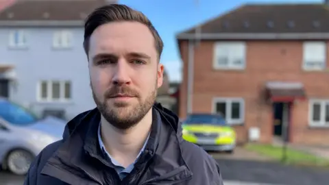 Kyle Moutray is standing in a street with a house behind him. A police car, which is parked outside the house, is out of focus. He has brown hair and a short beard. He is wearing a rain coat. 
