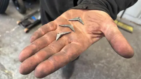 Three boomerang shaped pieces of metal which are about two inches long sit in the oily hand of a mechanic 
