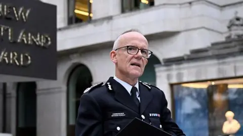 Reuters Sir Mark Rowley, pictured speaking at a podium outside the Met Police headquarters. A sign, partially cropped out of the picture, reads: New Scotland Yard. Mark Rowley is wearing glasses and has grey hair which is thin on top.