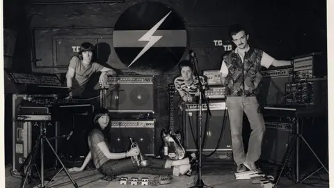Cosey Fanni Tutti Black and white picture of four musicians. There are three men and a woman who is sitting. They are surrounded by instruments with a logo behind them depicting a flash. The woman is holding a cornet.