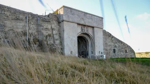 A general view picture of HMP The Verne in Portland, taken on 4 February 2022. The picture shows the entrance to a stone tunnel, with a coat of arms above it, and a stone wall on either side of it.