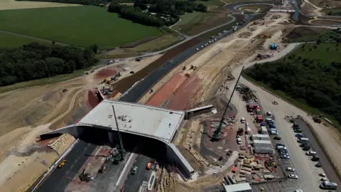 Aerial view of a major road construction project cutting through open countryside. In the foreground, a large concrete bridge spans a newly built road, with cranes, construction vehicles and building materials visible around the site. The partially completed road stretches into the distance towards a roundabout, while fields, woodland and temporary construction compounds surround the route. 