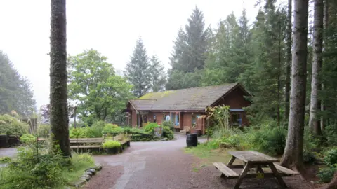 A wooden building, similar in design to a log cabin, surrounded by woodland.