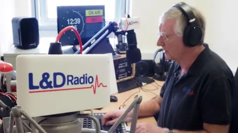 A man sits in a radio studio surrounded by speakers, a screen and a microphone. He is wearing headphones. There is L&D Radio branding