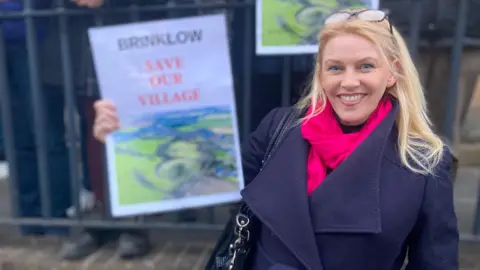 BBC Campaigner Linda Johnson holds a poster that says "Brinklow save our village". She is wearing a blue coat and pink scarf and has long blonde hair.