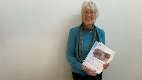 Parkinson's UK Dee Way standing against a wall smiling and holding a pamphlet