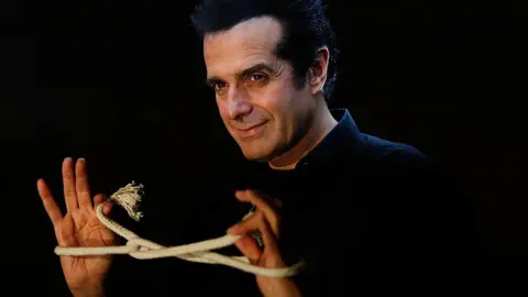 Getty Images Magician David Copperfield stands in front of a black background. He is wearing a black shirt, and is looking off to the side of the camera, smiling. He has a rope tied in an intricate knot around both hands, and is holding the ends.