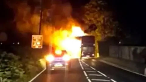 Island Echo A bus and car on the road - there are flames coming from the rear of the bus. it is night and the image is not completely clear.
