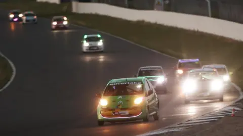 James Roberts Photography Silverstone Circuit at night with Citroen C1 cars of various colours taking a banked corner with their headlights shining. The front car is green, with a Silverlink banner across the top of the windscreen. There is a group of four cars close behind the leader.