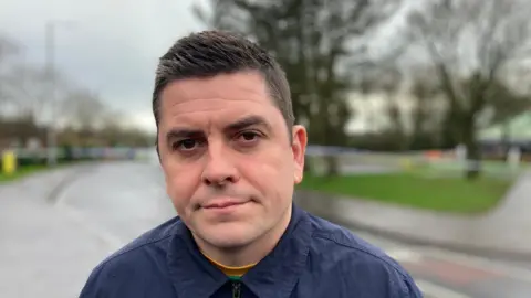 Sinn Féin MLA Danny Baker standing on a street infront of police cordons. He is wearing a dark blue shirt. He has short dark hair.