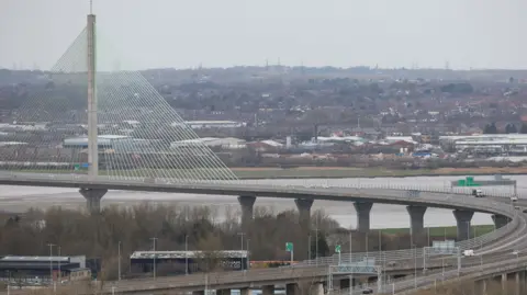 The Mersey Gateway toll bridge, which runs across the River Mersey. It is a cable-stayed bridge with the access road looping round from the west. 