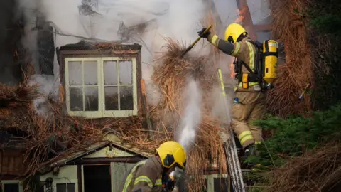 A firefighter stands on a ladder as they move burned straw from a thatched roof that is on fire. Smoke can be seen billowing from the property. Another firefighter is working on the ground underneath the one on the ladder.