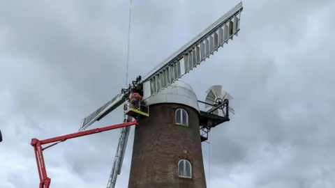 Sue Baxter Under a dark grey sky, the top of the windmill, which is a round red brick building with a white dome on top, has a cherry picker lifting men to the white sails as they remove one