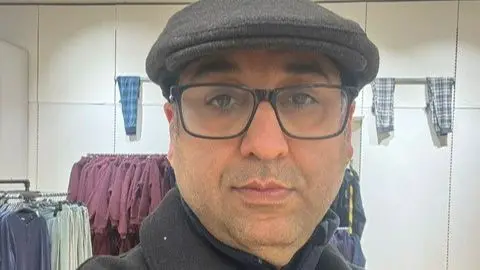 Hamidreza Bahmani Hamidreza Bahmani wearing a grey flat cap, dark framed glasses and a black coat. He is inside a clothes shop. 