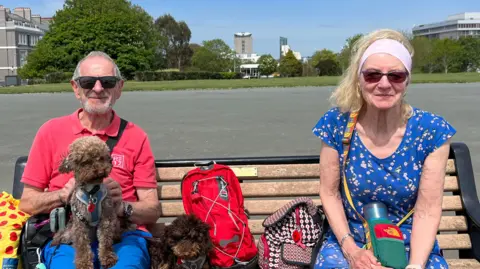 Bob and Rita Micek with their dogs Chico and Rolo sat on a bench on Plymouth Hoe. Rita is wearing a blue floral dress, and Bob is wearing a red t-shirt with blue shorts. One dog is sat on his lap, while the other one is sat beside him.