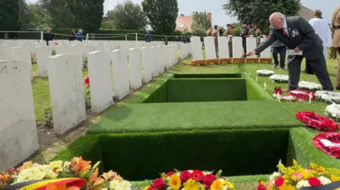 BBC/Olivia Richwald There is a whole in the ground which contains coffins. You cannot see the coffins. A man is leaning over the hole and throwing soil. There are colourful wreaths laid around the hole.