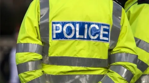 The back of a police officer wearing a hi-vis jacket, bright yellow-green with reflective silver strips and with a blue POLICE logo.
