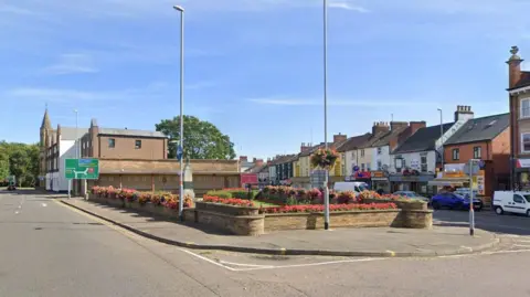 Google Abington Square in Northampton, showing a grass square bordered by a stone wall and flowers. There is a lamppost in front of the square on which a hanging basket is suspended. There are roads around the square, with two and three storey shops to the right of the road. A church is visible in the distance on the left.