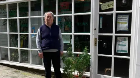Teresa Roberts Teresa Roberts stands in front of her shop wearing black trousers, a blue gilet and grey long-sleeved top, looking toward the camera with a plant to her side.