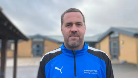 Shariqua Ahmed/BBC Scott Collins, a man with short, greyish hair and a beard, wearing a blue-and-black tracksuit top. It has a white puma logo and has Hampton Vale Primary Academy written on it on the right side.