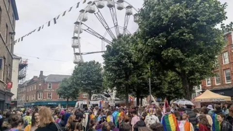 KIT TAYLOR/BBC Crowds of people in York holding a giant rainbow flag, with some holding flags and a big wheel ride is seen in the background.