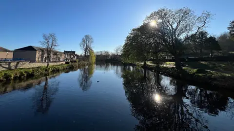 JulieJ View down a river in sunshine. On the left bank are trees in bud, reflected in the water, with the sun shining through the branches. To the left are a line of two-storey brick buildings, and three trees - two of them leafless - reflected in the water.