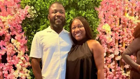 Daniel Udoeyo Daniel Udoeyo is wearing a white shirt and is standing with his arm around his wife Joy Kabiri who is wearing a brown dress. The couple are standing in front of a flower display of pink and white flowers.