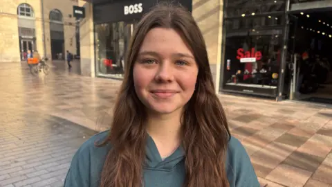 A brunette girl in a blue hoodie smiling at the camera 