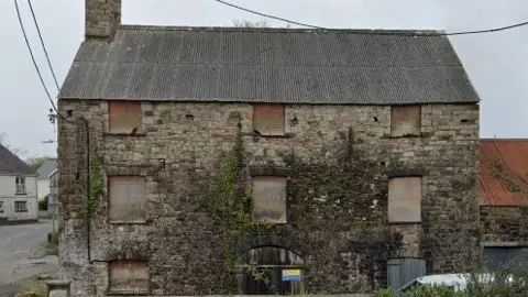 Google Old mill building with windows boarded up . Plants grow over part of the facade and some of the old stone has turned black. The wide wooden entrance door has a warning sign on it. 