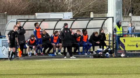 Prostate Cancer UK The image shows the technical area beside a football pitch. A row of substitutes and staff are seated or standing next to a covered dugout structure during a match.