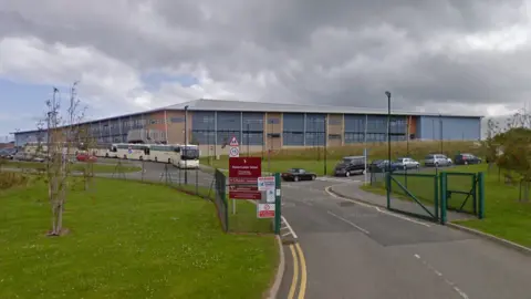 A large school building with a metal roof and multiple windows. Several vehicles, including buses and cars, are parked in front. A driveway with yellow lines leads to an entrance gate. A signpost is in the foreground.
