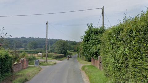 Swallowfield Road in Arborfield. It is a narrow county road with a hedge on the right of the road and a brick wall on the left. There are wires over the road from telegraph poles. A car and a motorbike are driving away from the perspective of the camera. 