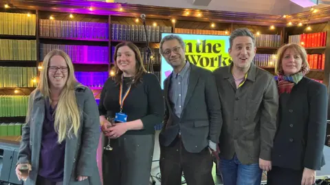 Jon Wright/BBC Poet Amy Wragg, Melissa Matthews, from Suffolk Community Libraries, Robin Deacon, artistic director of Spill, and Douglas Rintoul and Sally Jones from New Wolsey Theatre stand in a line in front of a stage.