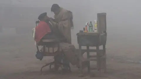 AFP A Pakistani barber shaves a customer alongside a road amid heavy fog and smog conditions in Lahore on January 24, 2019.