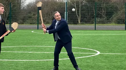 BBC Paul Givan playing hurling with a pupil on a sports field. Both are holding hurling sticks - Givan is tossing a sliotar in the air and preparing to hit it. He is wearing a dark blue suit, light blue shirt and sliver tie.