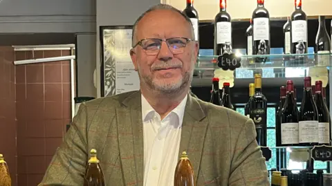 Pub owner Nigel Farnfield behind the bar at The Olive Tree in Sutton Green, pictured with ale pumps and a wall of wine bottles.