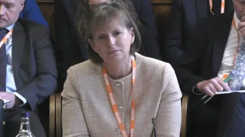 UK Parliament Heidi Mottram. She has a bob haircut and white buttoned cardigan. She is wearing an orange visitors lanyard. 