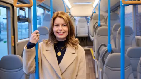 NECA Kim McGuinness in brown coat standing in a bus posing for the camera. She is smiling and holding a blue pole.