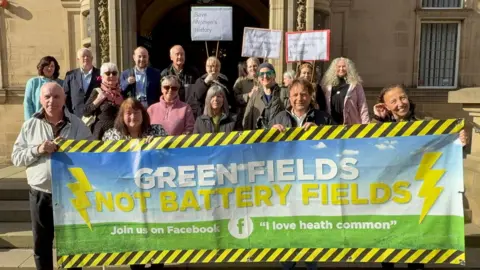 LDRS A mixed gender group of people stand in front of a large building, holding a banner between them. The banner reads 'Green fields not battery fields'.
