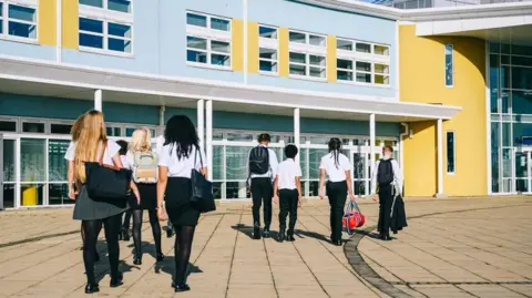 Pupils in school uniform walk towards a school building in the sun. 