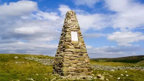 A pyramid shaped stone cairn on the top of a Borders hillside. There are stones littered around it on a grassy landscape with a cloudy blue sky above.