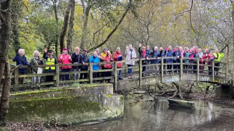 A large group of walkers, wearing different coloured coats, standing on wooden footbridge over the River Nadder. 