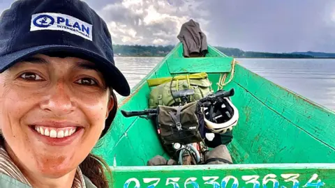 Ellie Mitchell-Heggs Mitchell-Heggs on a bright green canoe on the Zambezi river. She is wearing a navy cap with the logo for Plan International on it. She is smiling at the camera and you can see her bike in the canoe with her helmet and the river behind her. There is green foliage in the far distance.