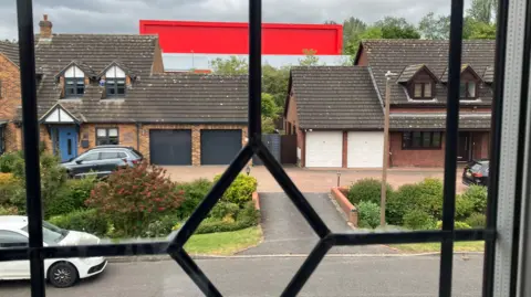 Amy Holmes/BBC A view through a window with black leaded bars looks out onto a quiet residential street with brick houses, driveways, and parked cars, with a large red industrial building visible beyond under a cloudy sky.