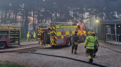 A clearing in a forested area, with a wooden building to the right. There are two fire engines with hose reels leading off camera. Firefighters walk about the scene.