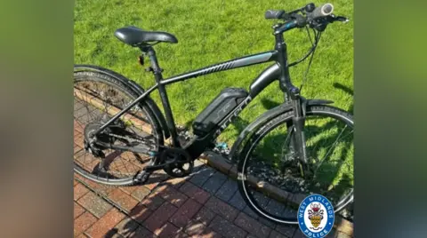 WMP handout The e-bike is stood on a hard brick ground next to some grass in the sunshine. It is black with some grey markings.