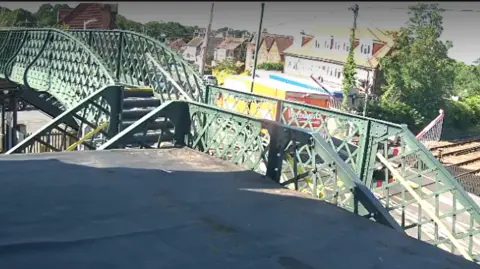 CCTV from GTR shows a green metal pedestrian footbridge overlooking a level crossing in Billingshurst, with a lorry visible beyond railway barriers and tracks below in bright sunlight.